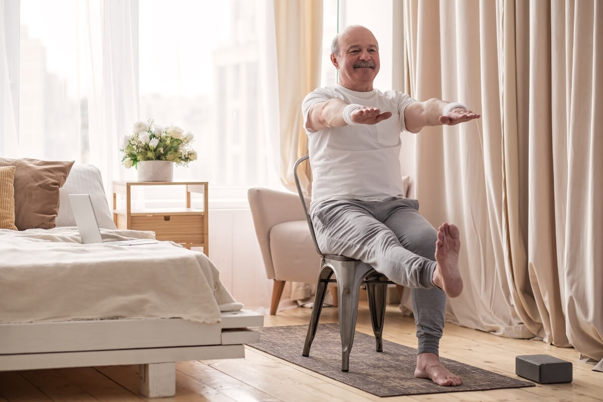 An older man smiling while seated on a metal chair in a light-filled bedroom, performing a single-leg extension exercise.