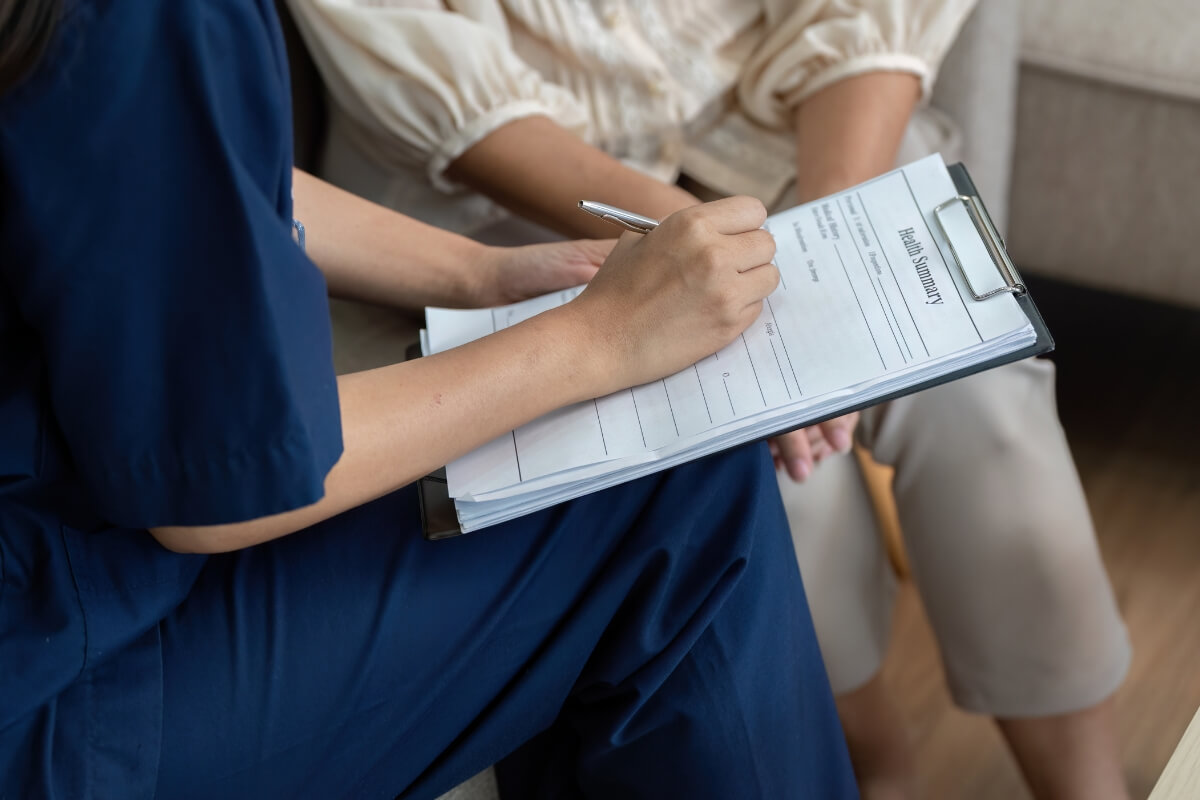Close-up of a person in navy scrubs writing on a clipboard labeled "Health Summary" while conducting a home assessment.