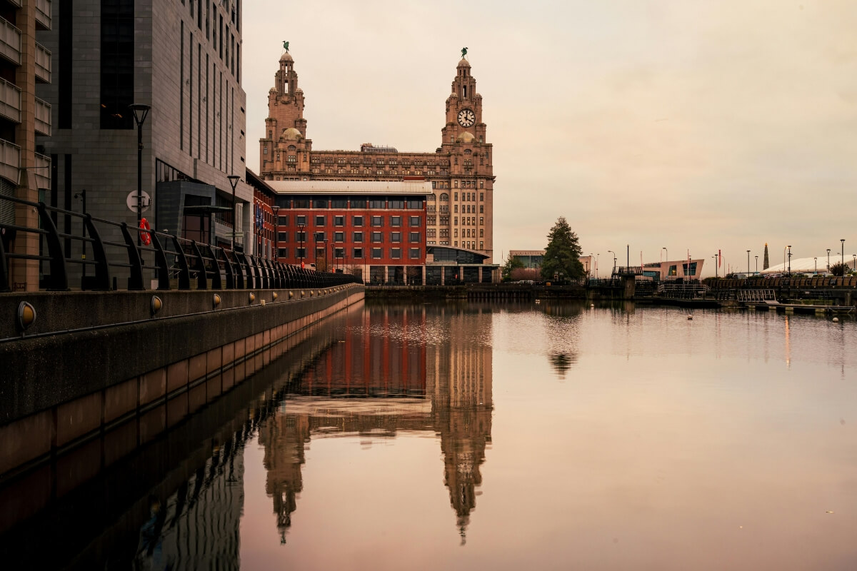 The Royal Liver Building in Liverpool, with its two clock towers, reflected in the calm water of the docks at sunset, flanked by modern buildings on the left.