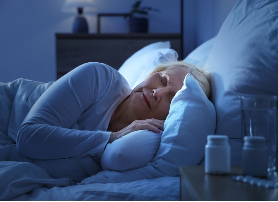 An older woman sleeping peacefully in bed with pill bottles and a glass of water on the nightstand.