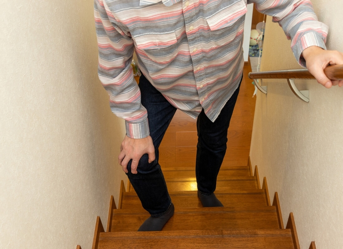 A person clutching their knee in discomfort while holding onto a handrail on a wooden staircase.