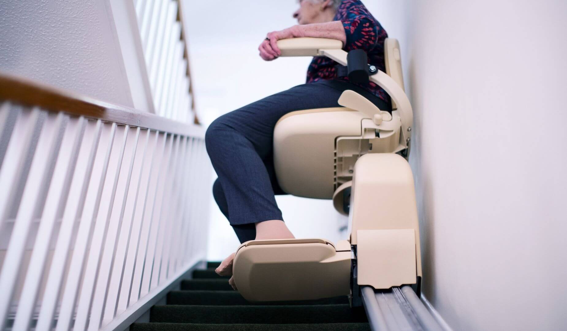  A low-angle view of a person's torso and legs, sitting on a stairlift as it moves up a carpeted staircase.