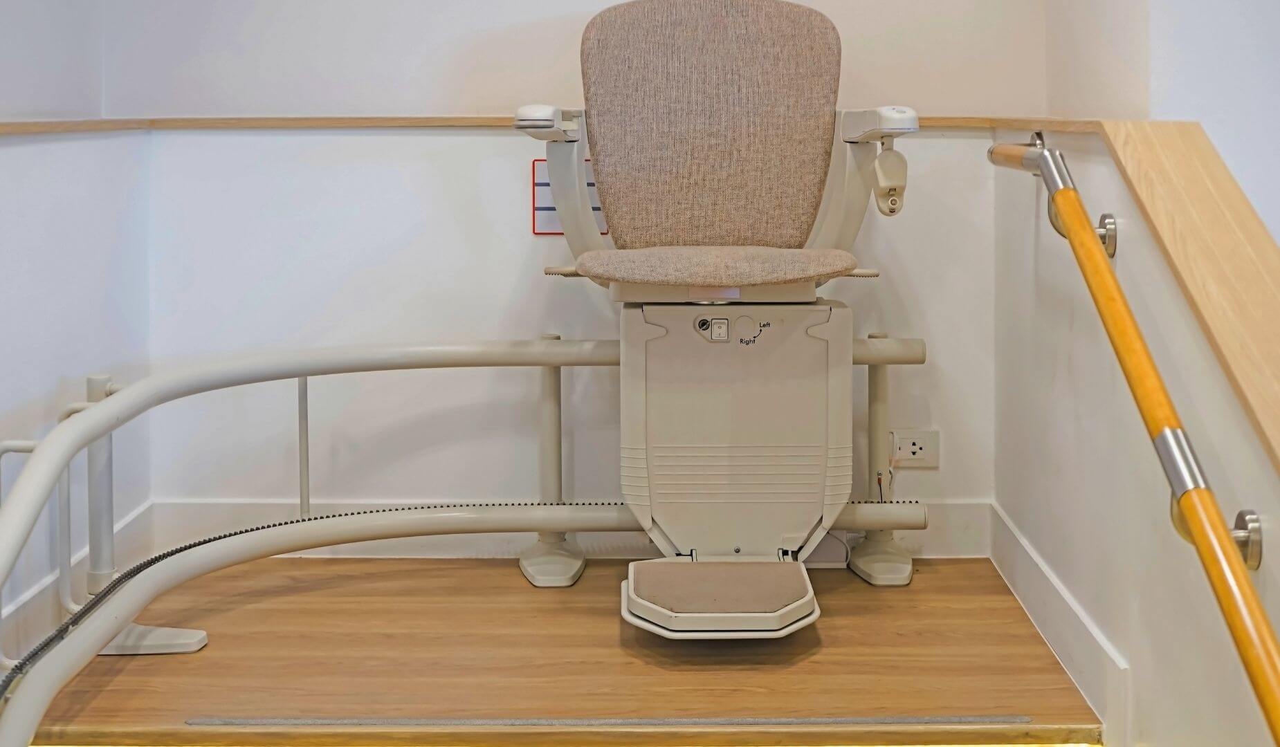 A close-up of a stairlift chair with a textured seat, parked at the top of a staircase with a wooden handrail.