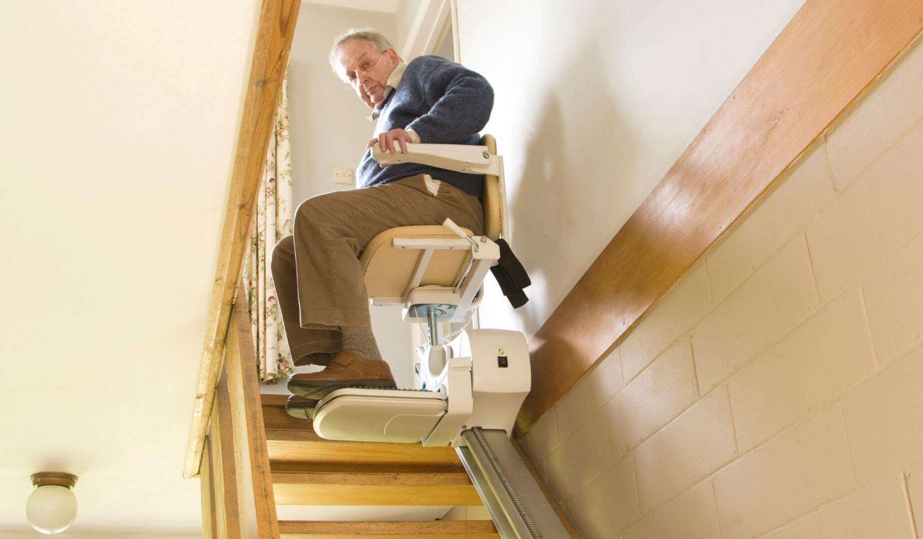A low-angle shot of an elderly person sitting on a stairlift, holding the armrests, as the lift travels up a wooden staircase.
