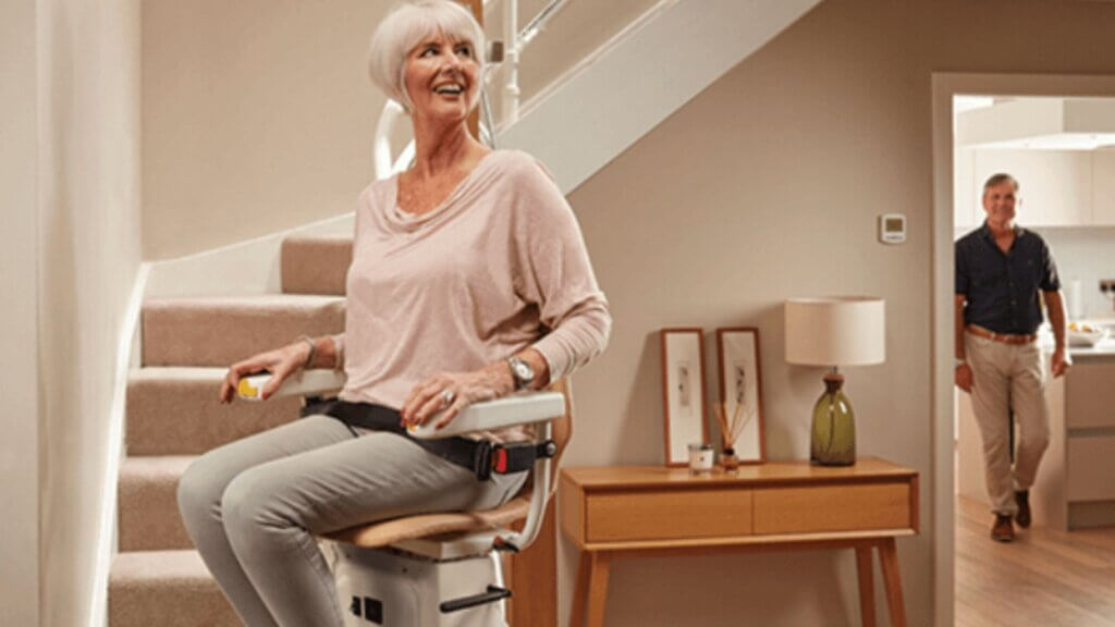 A smiling elderly woman riding a stairlift up the stairs while a man walks through a doorway in the background.