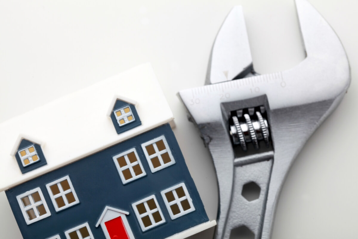 A small, miniature house model (blue and white) is placed next to a large, shiny wrench. This represents home maintenance, modification and safety for independent living.