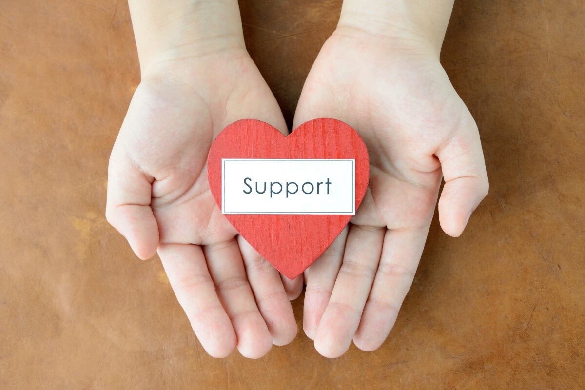A close-up of two cupped hands holding a red, heart-shaped cutout with a white label that reads "Support" in the centre. This symbolises community or personal care and emotional support.