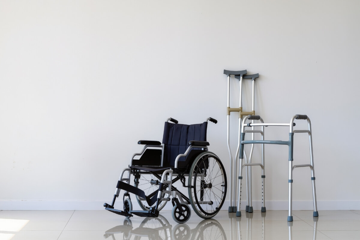 A clean, brightly lit room with a white wall features various mobility aids: a wheelchair, a pair of crutches and a walker. This provides a visual representation of available mobility equipment.