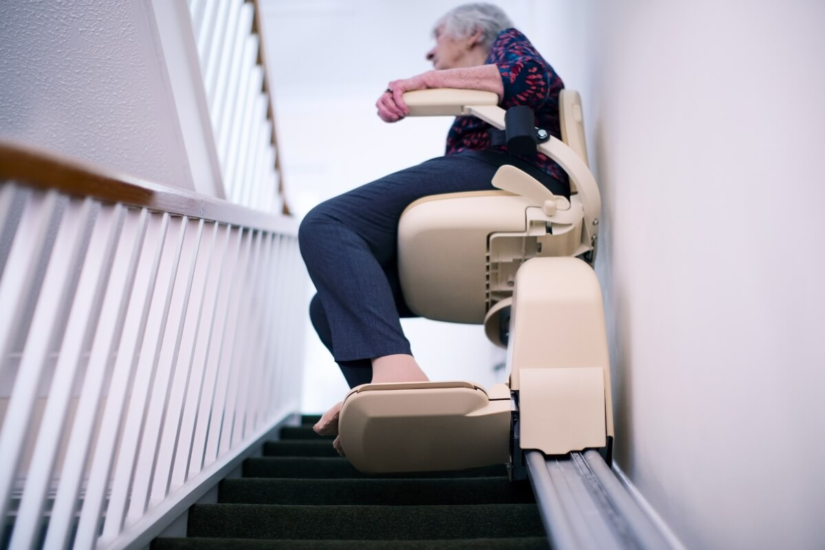 A low-angle shot of an older woman using a stair lift/chair lift as it ascends a carpeted staircase. This reinforces the theme of using specialised equipment to maintain access to all areas of the home.