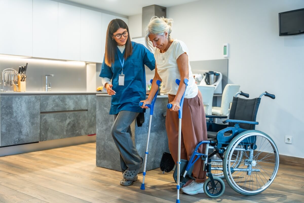 A healthcare provider (wearing blue scrubs) is assisting an older woman who is using crutches. An empty wheelchair is nearby. They appear to be in a modern kitchen/living area, suggesting home care or physical therapy. This illustrates physical assistance and rehabilitation.