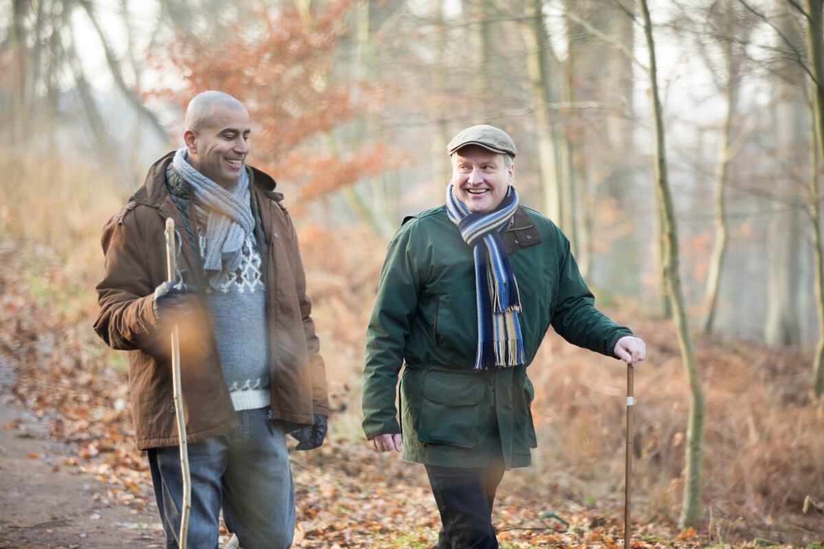Two men in winter coats walking through a wooded path with walking sticks.
