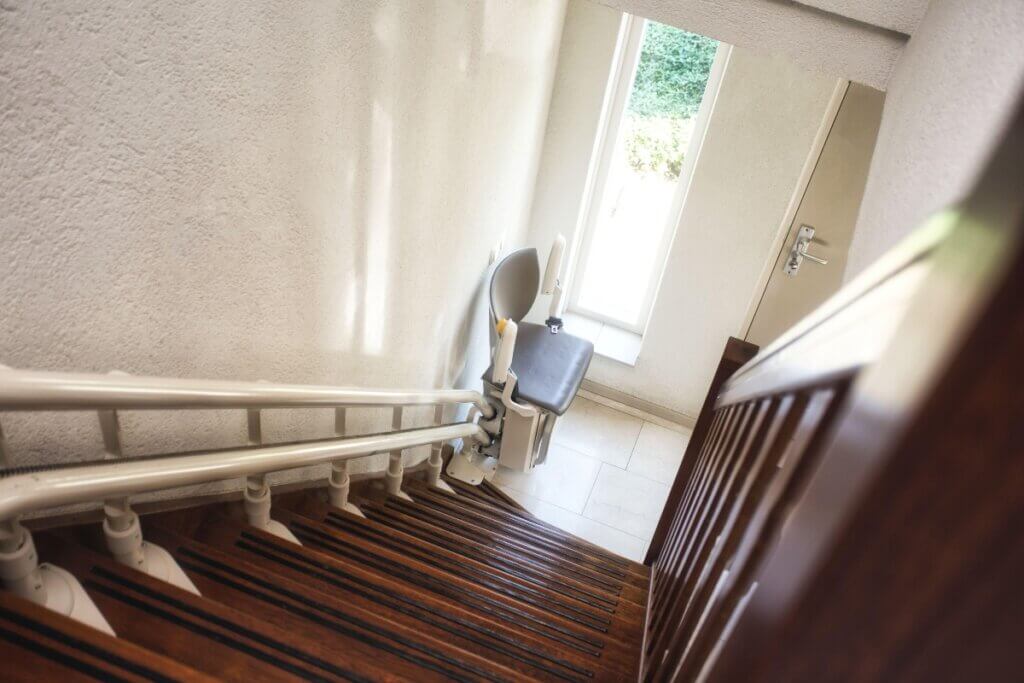 A high-angle view looking down a wooden staircase showing a stairlift unit mounted on a double-rail system near a doorway.