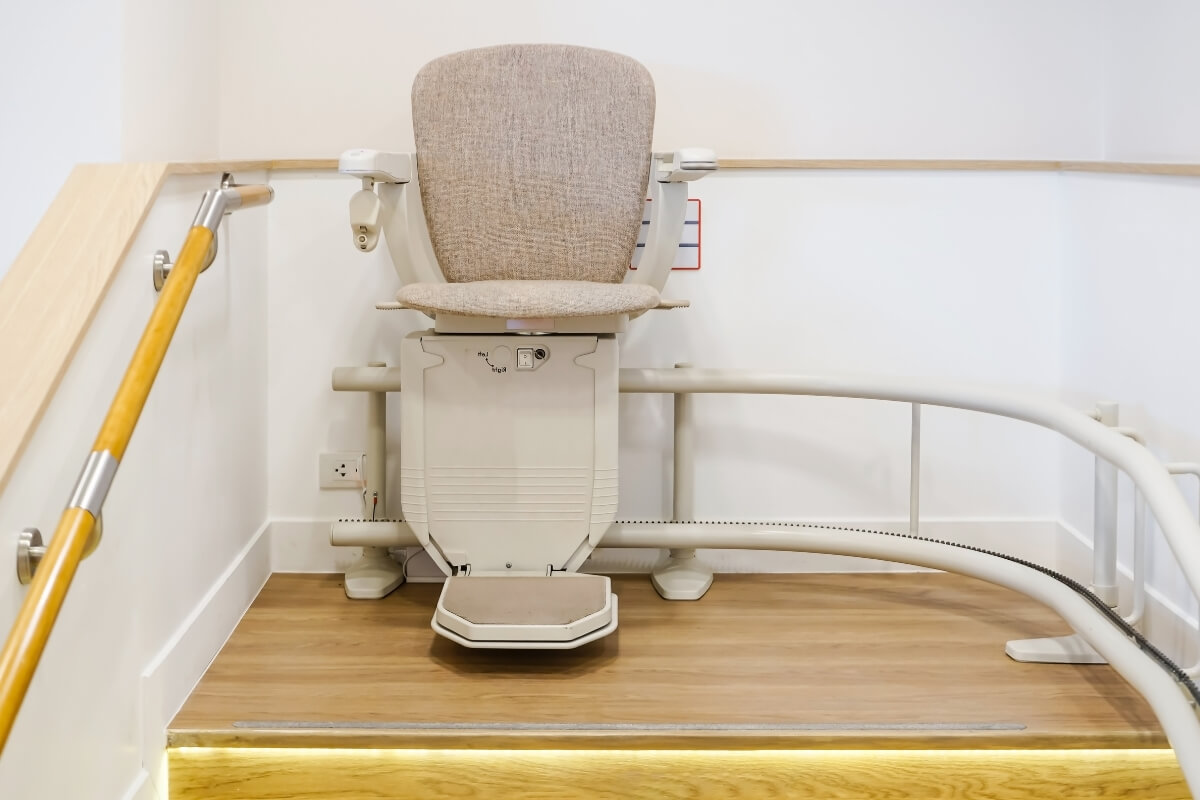 A beige fabric-upholstered stairlift parked at the top of a curved rail system on a wooden landing.