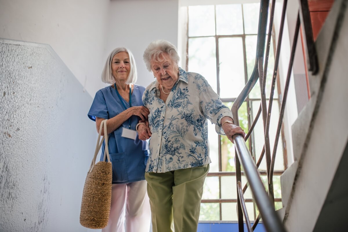 A healthcare worker assists an older adult in walking down a set of stairs while holding the handrail.
