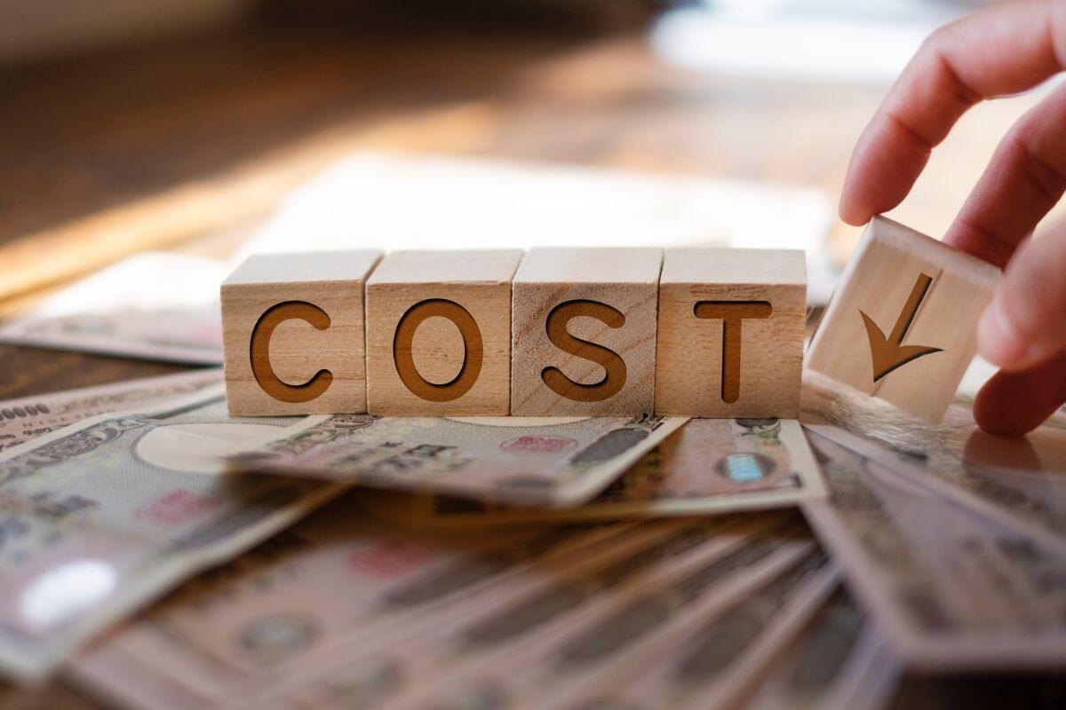 Wooden blocks spelling out the word "COST" sitting on top of Japanese Yen banknotes, with a hand placing a final block featuring a downward-pointing arrow next to them.