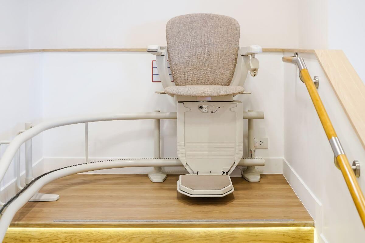 A neutral-toned stairlift with a textured beige seat parked on a curved white rail at the top of a wooden staircase.