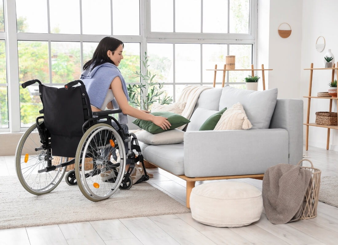 A woman in a wheelchair adjusting a green pillow on a light grey sofa in a bright living room.