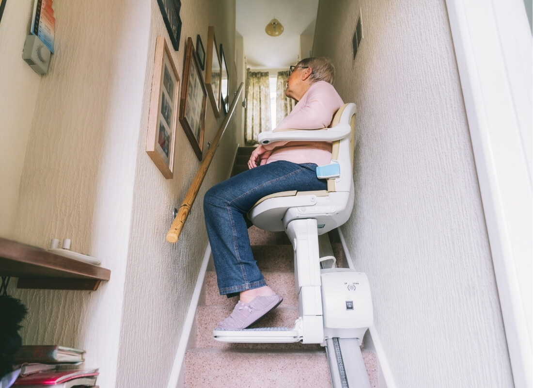 An elderly person sitting on a motorized stairlift while ascending a carpeted staircase in a home.