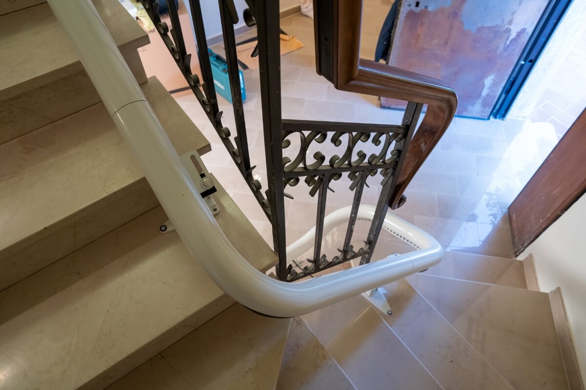 A high-angle view of a curved white stairlift rail installed along a marble staircase with decorative metal bannisters.