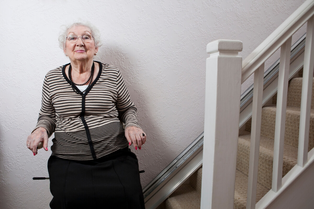 An elderly woman sitting comfortably on a stairlift positioned at the bottom of a carpeted home staircase with white railings.