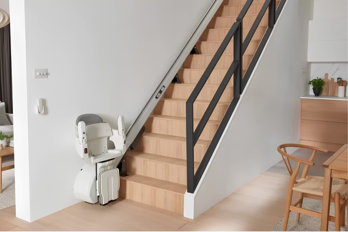 A modern white stairlift parked at the bottom of a wooden staircase in a bright home interior.