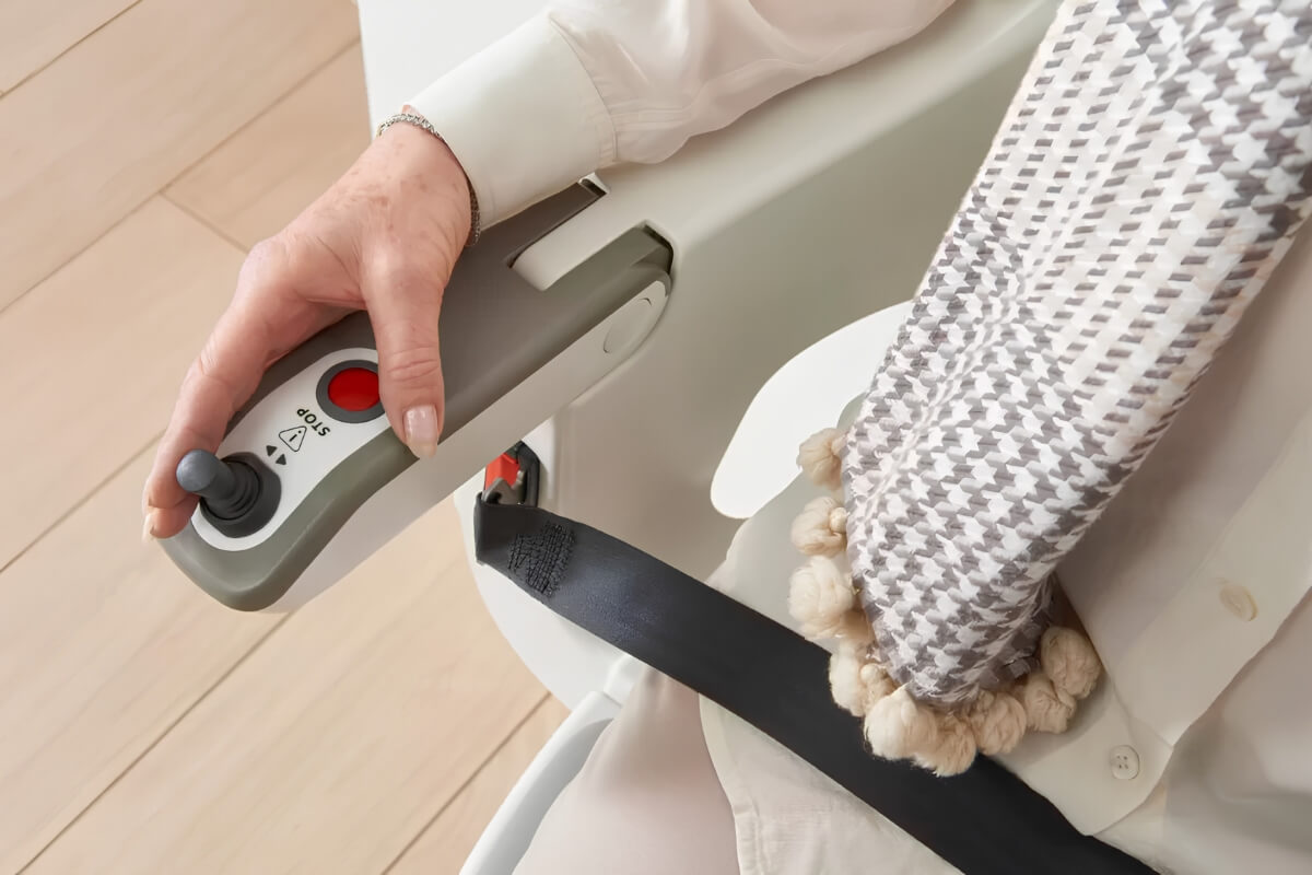 A close-up of a person's hand using a joystick controller on the armrest of a stairlift.