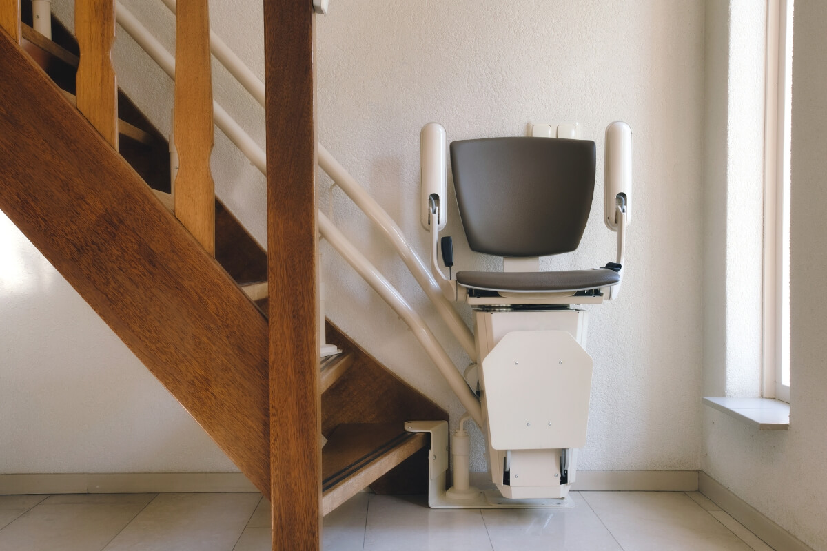 A modern white stairlift chair with its seat folded up, parked neatly out of the way at the top of a carpeted landing next to a wooden banister.