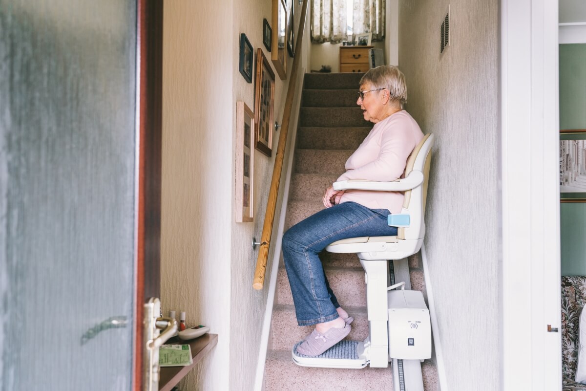 An elderly woman sitting on a white and cream motorised stairlift, riding up a narrow, carpeted staircase inside a home.