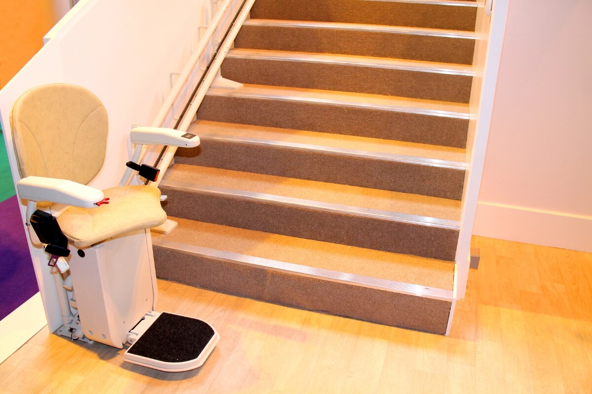 A cream-coloured stairlift parked at the base of a carpeted wooden staircase with a silver metal track running along the side wall.