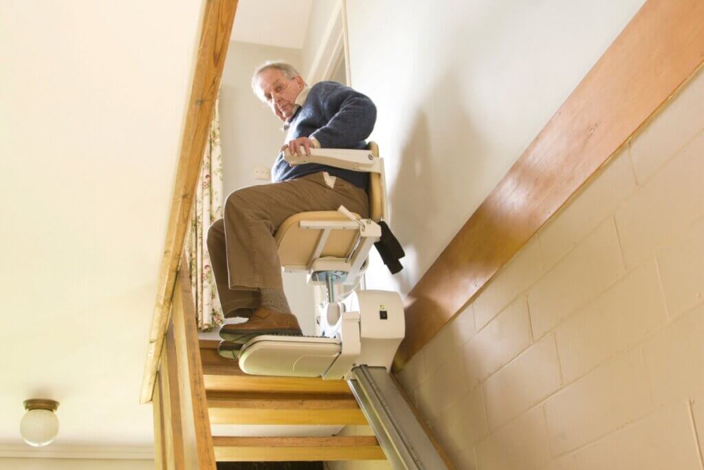 A low-angle view looking up a wooden staircase at an elderly man using a motorised stairlift to ride up to the second floor of a home.