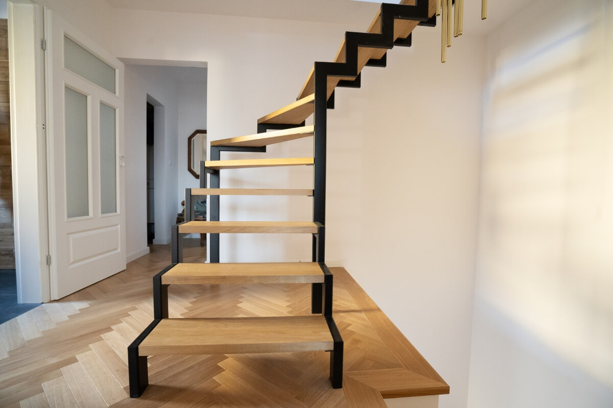 A cream-coloured stairlift parked at the bottom of a straight wooden staircase with a grey carpet runner.