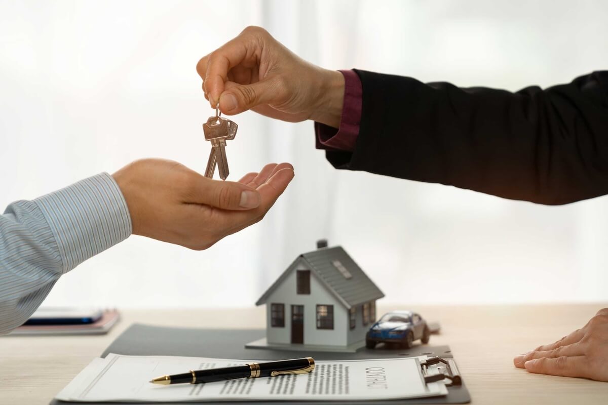 A close-up shot of a person in a dark suit handing a set of keys to another person. In the background on the desk, there is a miniature model house, a small blue toy car, a pen, and a signed contract.