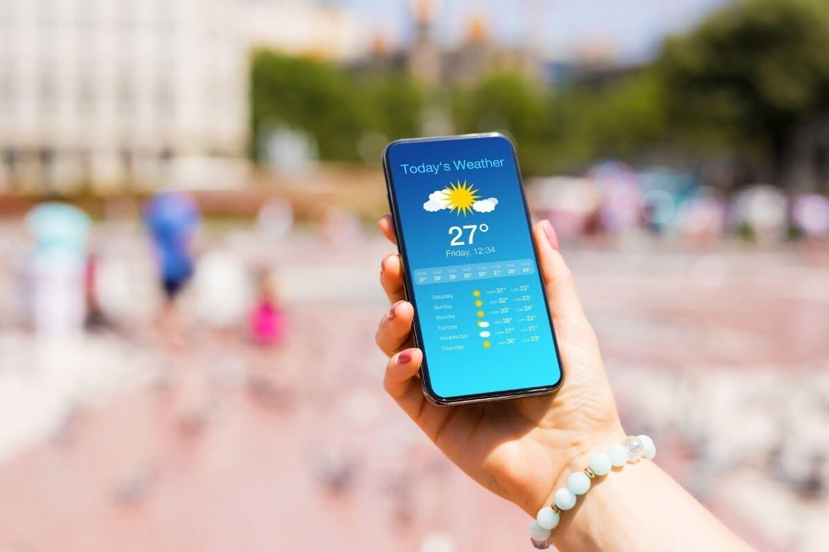 A person holding a smartphone outdoors displaying a sunny weather forecast app screen showing 27°C.