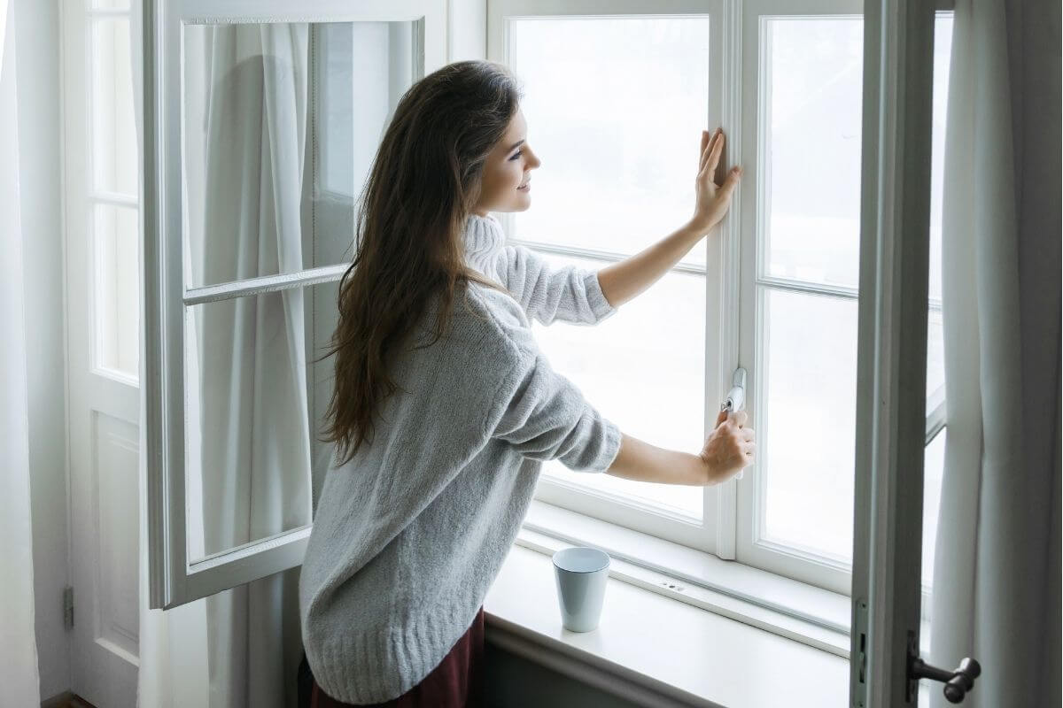A woman wearing a knit sweater standing by an open window to let fresh air into a room.