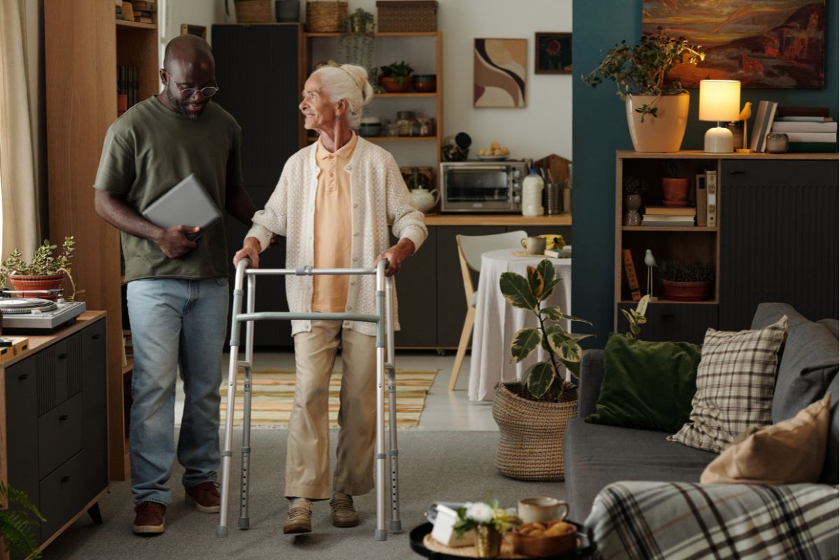 A man walking attentively beside an elderly woman who is using a metal walking frame indoors.