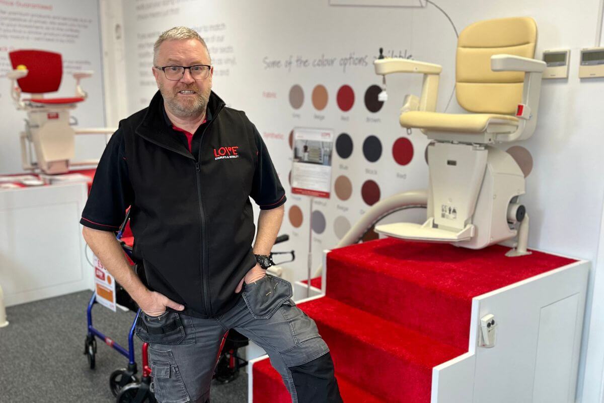 A man in a work jacket standing inside a showroom next to a stairlift demo unit mounted on red stairs.