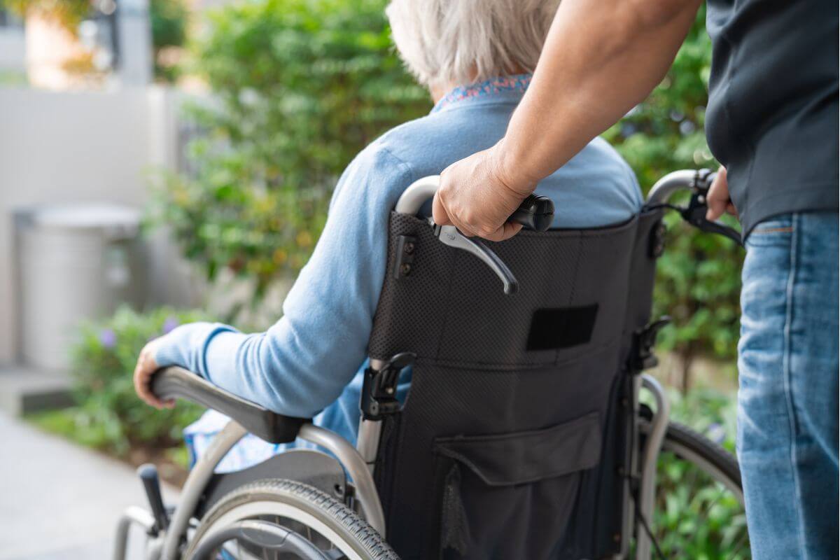 Rear view of a person pushing an elderly individual sitting in a wheelchair outdoors.