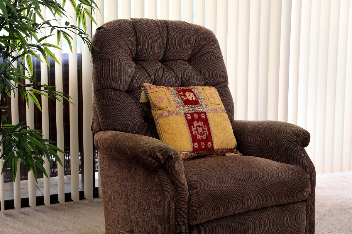 A plush brown corduroy armchair with a patterned yellow and red decorative pillow set in front of vertical window blinds.